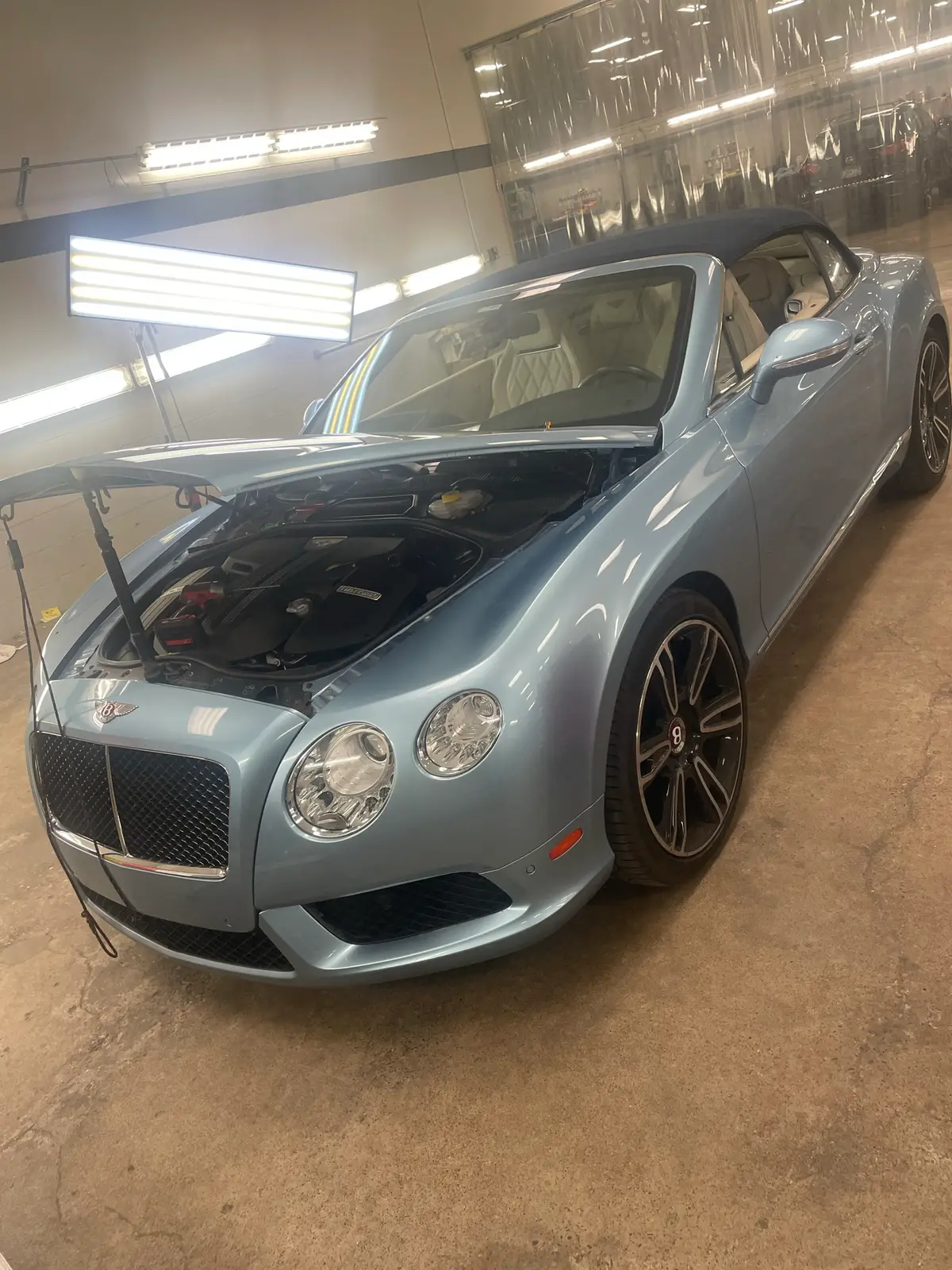 Light-blue Bentley Continental convertible in the Hail Solutions shop bay under LED lighting